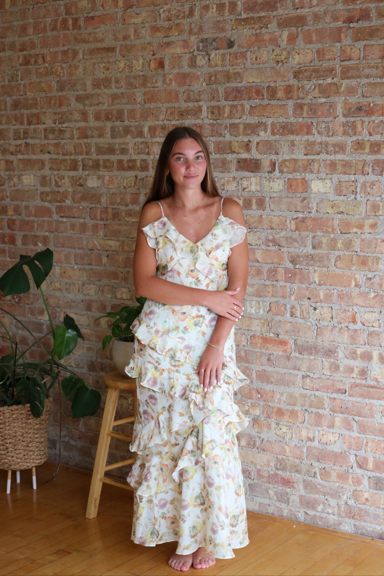 Woman in a floral dress standing against a brick wall with a plant in the background