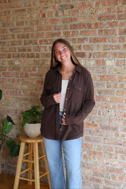 Woman wearing a brown jacket standing against a brick wall with a plant and wooden stool in the foreground.